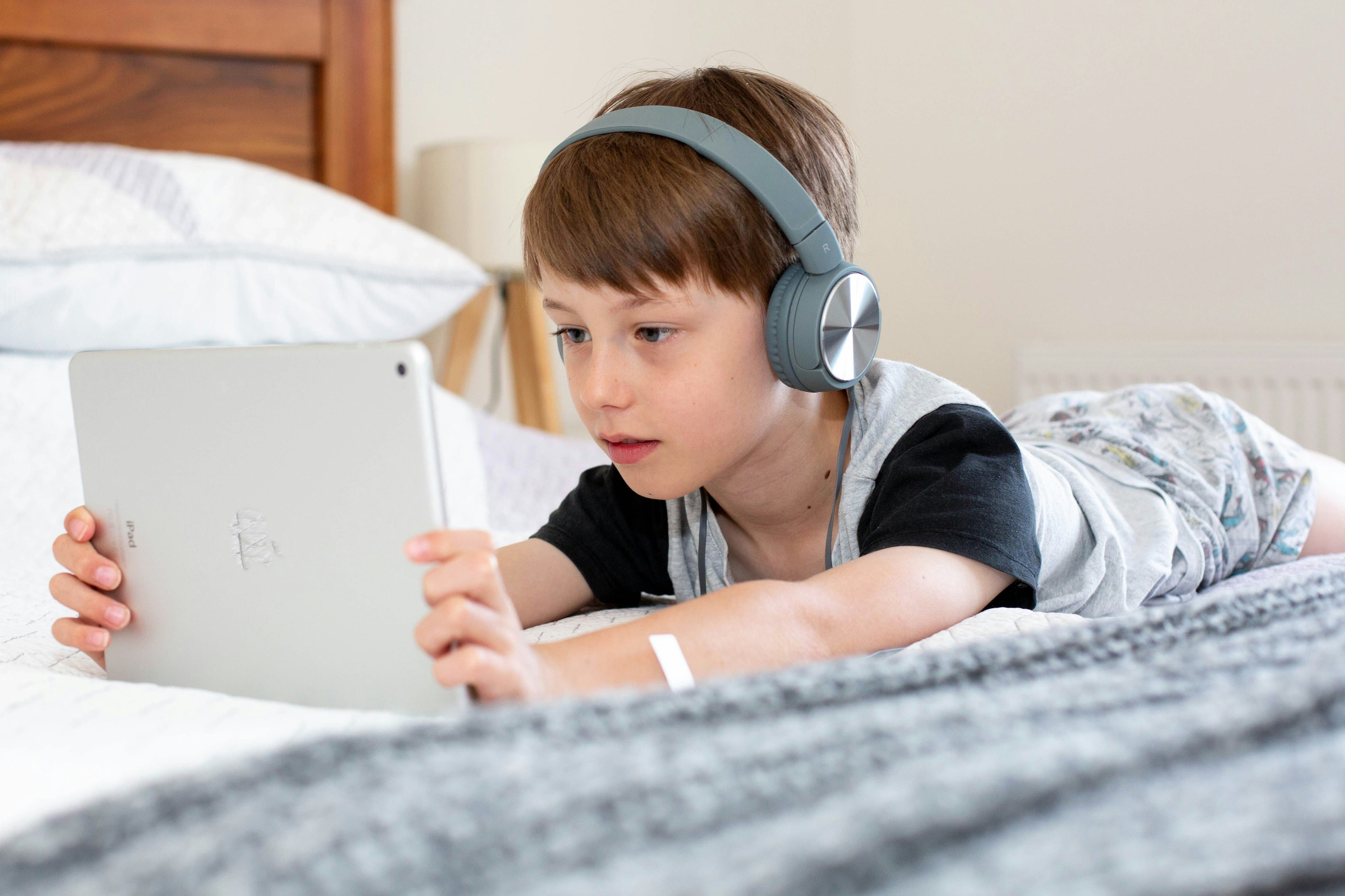 Jonge jongen liggend op bed met een tablet in zijn handen en een hoofdtelefoon op, geconcentreerd kijkend naar het scherm.